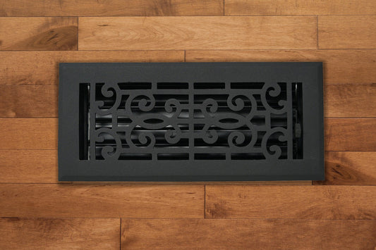 The Madelyn Carter Cast Aluminum Baroque Vent Cover in black, shown on a light brown wooden floor, adds elegance and airflow control with its scroll design and adjustable metal dampers.