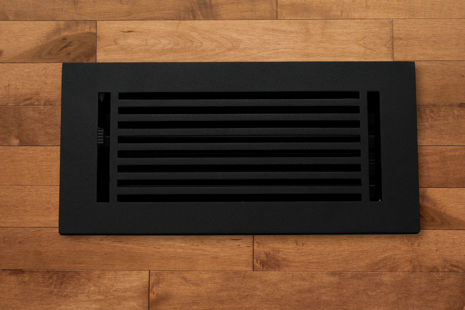 The Madelyn Carter Cast Aluminum Linear Bar Vent Cover in black, featuring horizontal slats, is installed in a light brown hardwood floor for a sleek and durable finish.