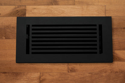 The Madelyn Carter Cast Aluminum Linear Bar Vent Cover in black, featuring horizontal slats, is installed in a light brown hardwood floor for a sleek and durable finish.