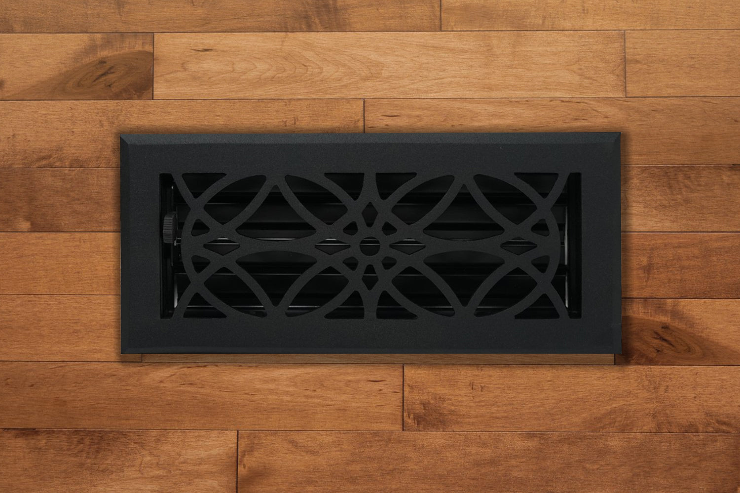 The Madelyn Carter Cast Aluminum Empire Vent Cover in black features a decorative geometric pattern, adding a stylish touch to light brown wood flooring and enhancing the elegance of any space.
