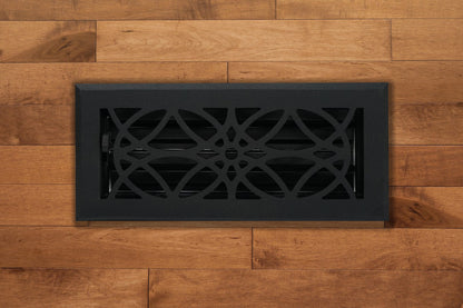 The Madelyn Carter Cast Aluminum Empire Vent Cover in black features a decorative geometric pattern, adding a stylish touch to light brown wood flooring and enhancing the elegance of any space.