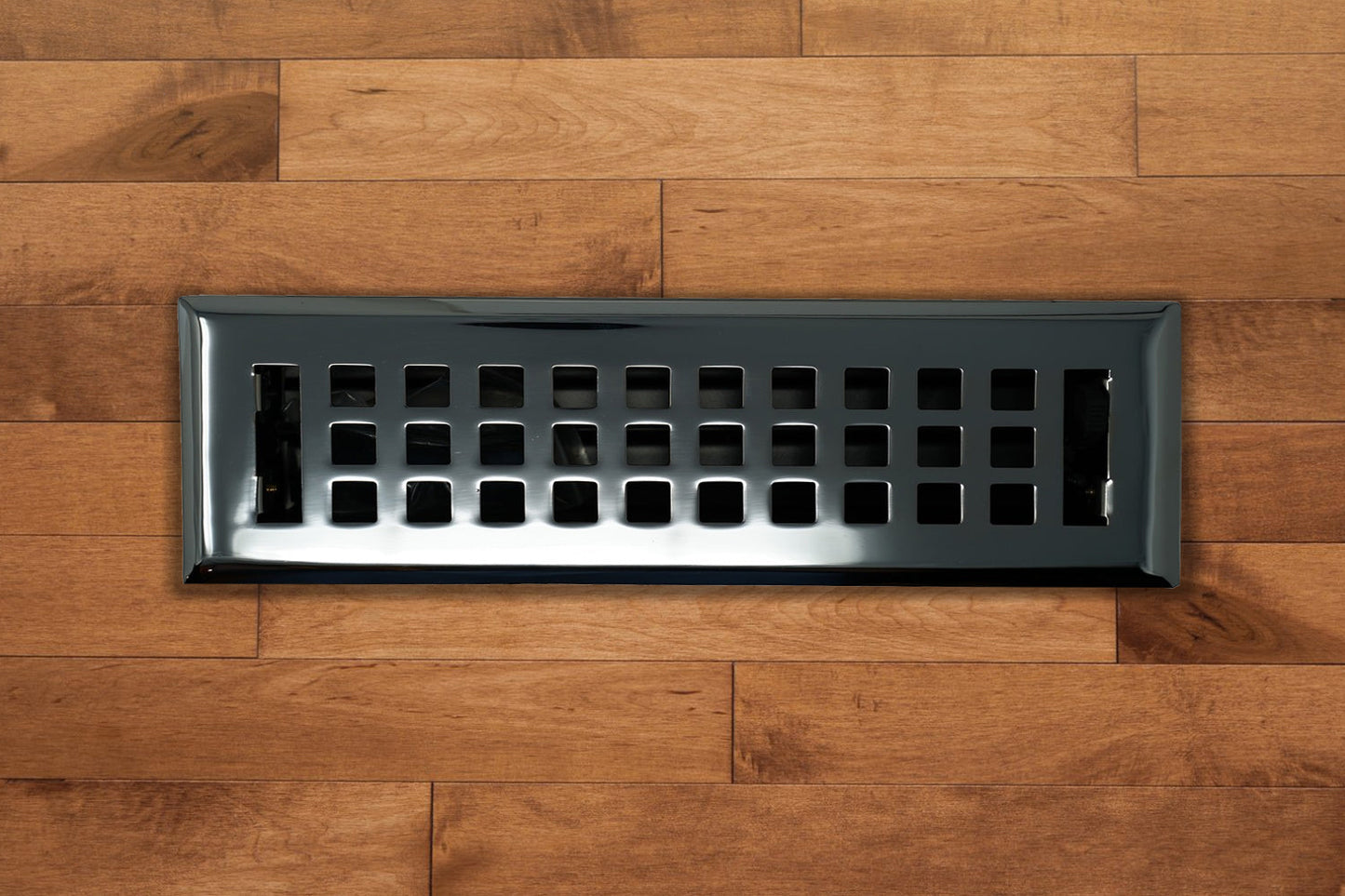 A Madelyn Carter Cast Brass Artisan Vent Cover in Polished Chrome with square openings is installed in a wooden floor with a medium brown finish.