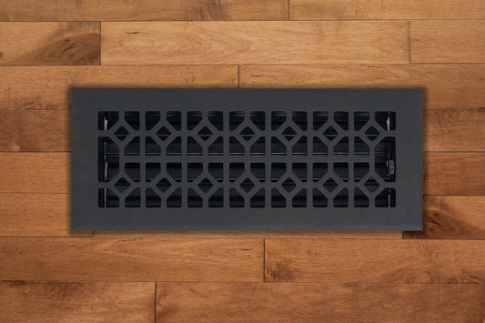 The Madelyn Carter Cast Aluminum Roman Vent Cover in black features a geometric pattern and is installed in medium-tone wood flooring, bringing a modern touch to the space.