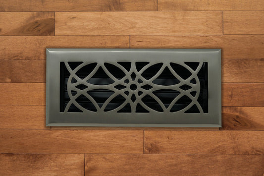 The Madelyn Carter Cast Brass Empire Vent Cover in Brushed Nickel features an ornate, symmetrical cutout design and is shown set in light brown wooden flooring.