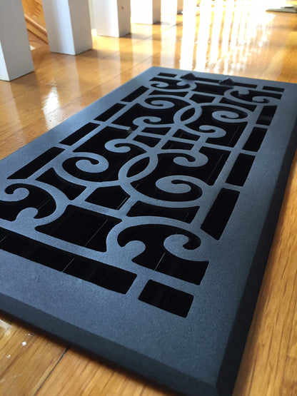 A black Madelyn Carter Cast Aluminum Baroque Vent Cover with a scroll pattern sits on polished wood flooring, sunlight streaming in the background.