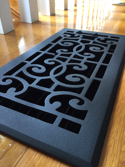 A black Madelyn Carter Cast Aluminum Baroque Vent Cover with a scroll pattern sits on polished wood flooring, sunlight streaming in the background.