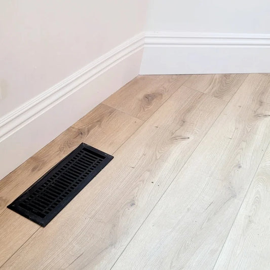 The Madelyn Carter Steel Modern Vent Cover - Black is installed in light wood flooring near a white baseboard and wall corner, adding a modern touch.