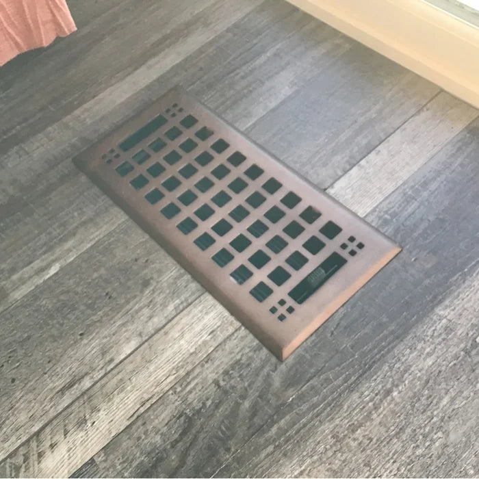 The Madelyn Carter Steel Artisan Vent Cover in Oil Rubbed Bronze features a grid pattern and is seamlessly installed in hardwood flooring near a window, providing both durability and style.
