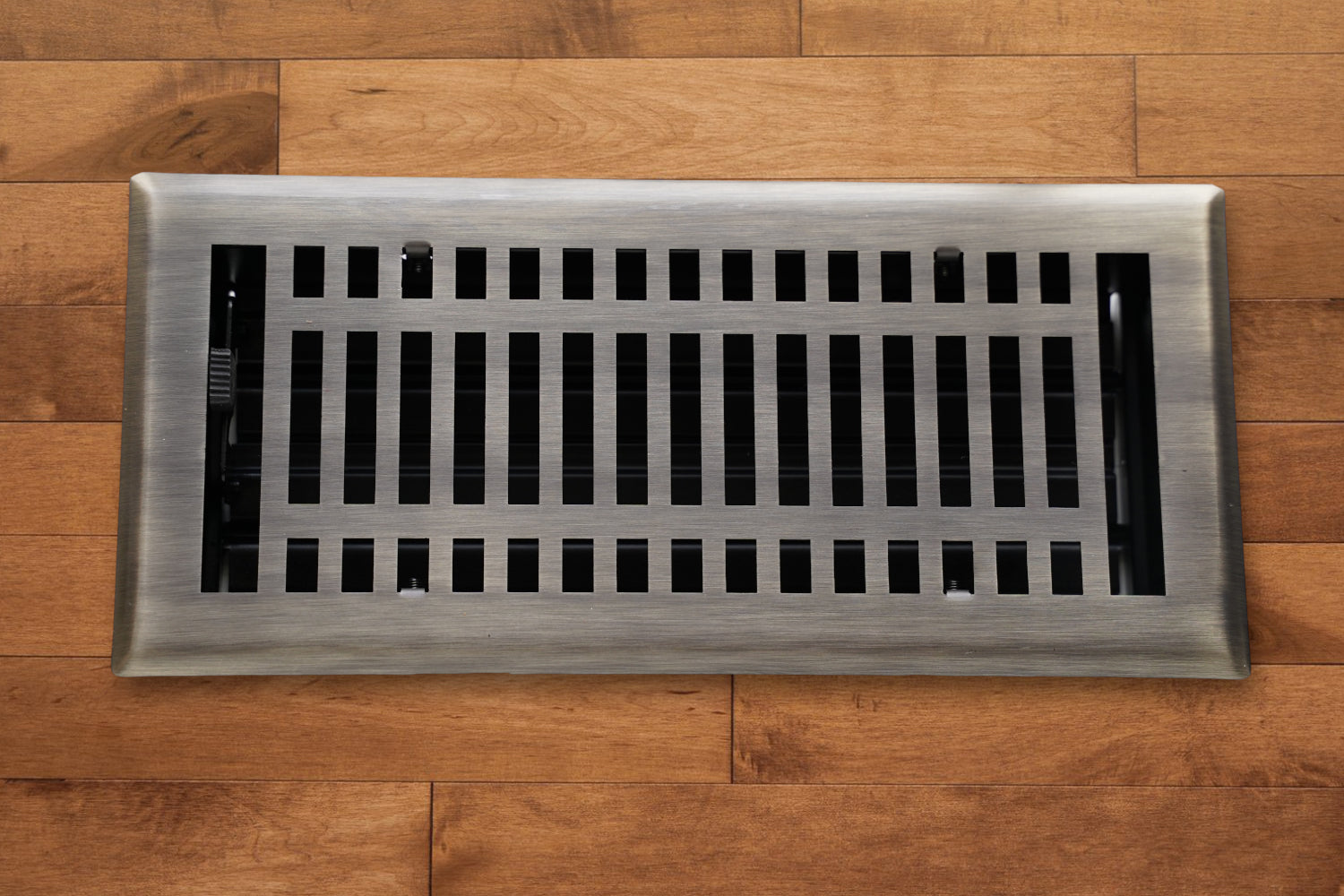 A Madelyn Carter Steel Modern Vent Cover in Antique Brass with a grid pattern is installed on light brown wooden flooring.