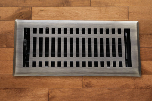 A Madelyn Carter Steel Modern Vent Cover in Antique Brass with a grid pattern is installed on light brown wooden flooring.