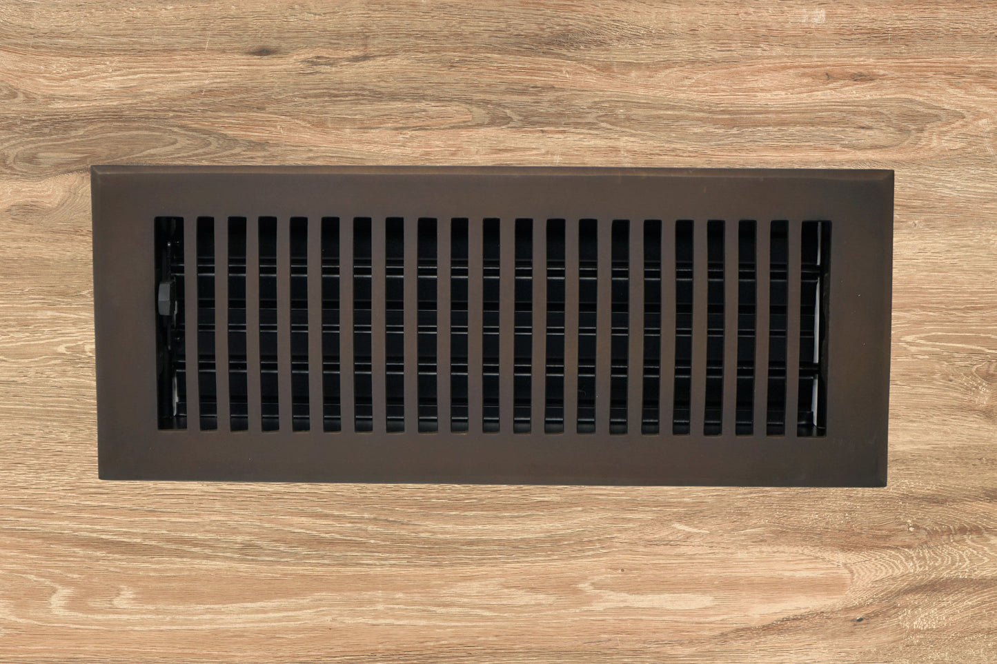 The Madelyn Carter Cast Brass Contemporary Vent Cover in oil rubbed bronze is installed in a wooden floor, offering a modern upgrade to traditional registers.
