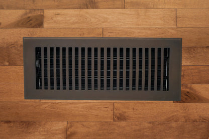 The Madelyn Carter Cast Brass Contemporary Vent Cover in Oil Rubbed Bronze is installed on a light brown wooden floor, offering a modern blend of style and function for air vent covers.