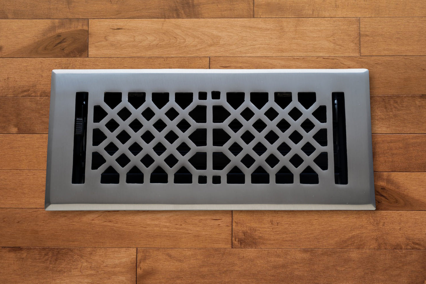 The Madelyn Carter Cast Brass Vintage Industrial Vent Cover in Brushed Nickel features a geometric pattern, adding elegance to any wooden floor.