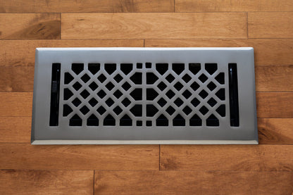 The Madelyn Carter Cast Brass Vintage Industrial Vent Cover in Brushed Nickel features a geometric pattern, adding elegance to any wooden floor.