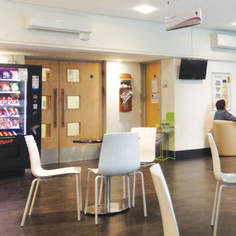 A waiting area with white chairs, a vending machine, wall-mounted TV, and a person near a notice board features the Aspen Mini White 100-250v Condensate Pump for its high wall indoor unit.