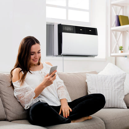 A woman sits on a couch using her smartphone in a living room with a Friedrich Kühl® Window Smart Wi-Fi Room Air Conditioner installed behind her.
