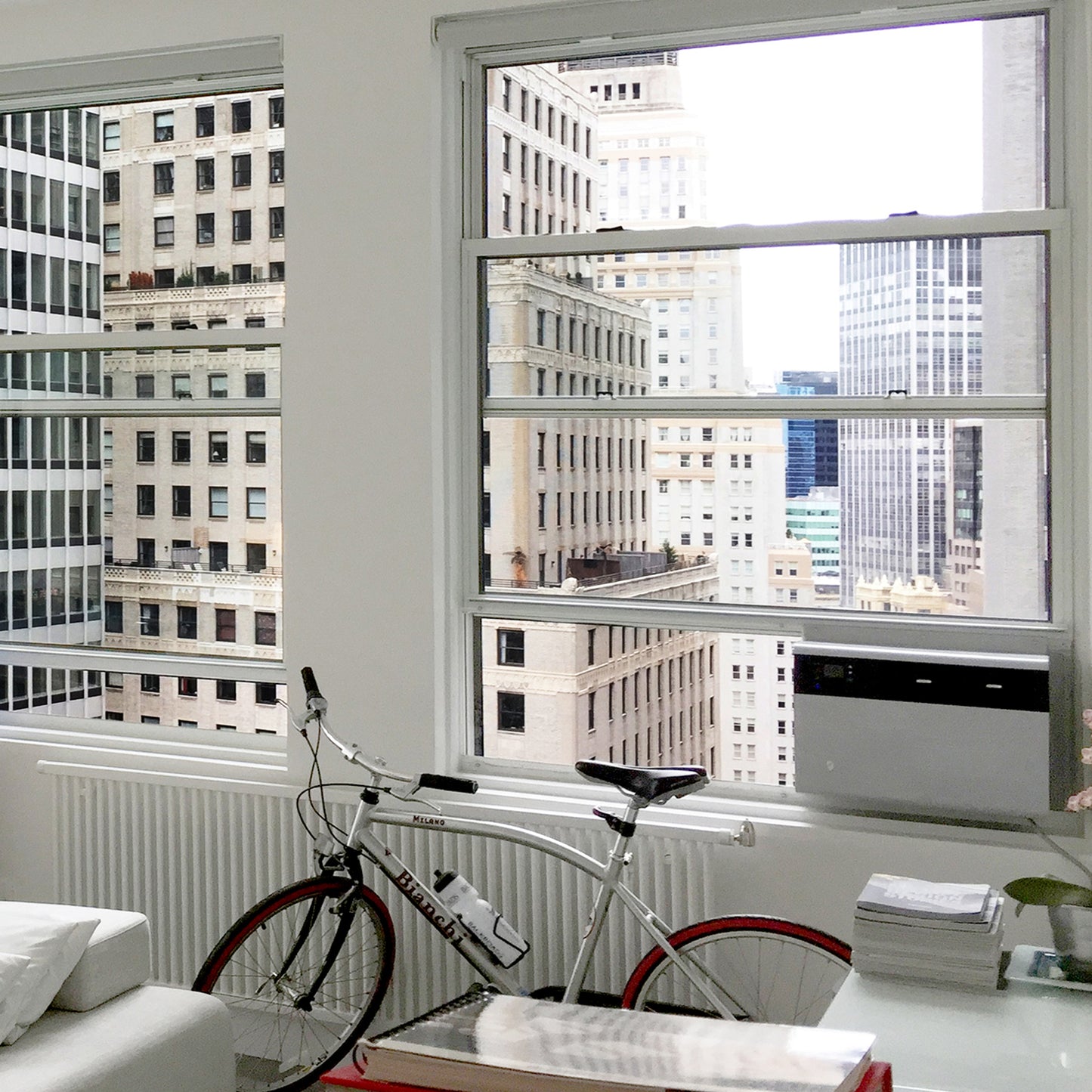 A white bicycle is parked near large windows in a modern apartment with city skyscrapers outside. A Friedrich Kühl® Window Smart Wi-Fi Room Air Conditioner by Friedrich is installed in one window.