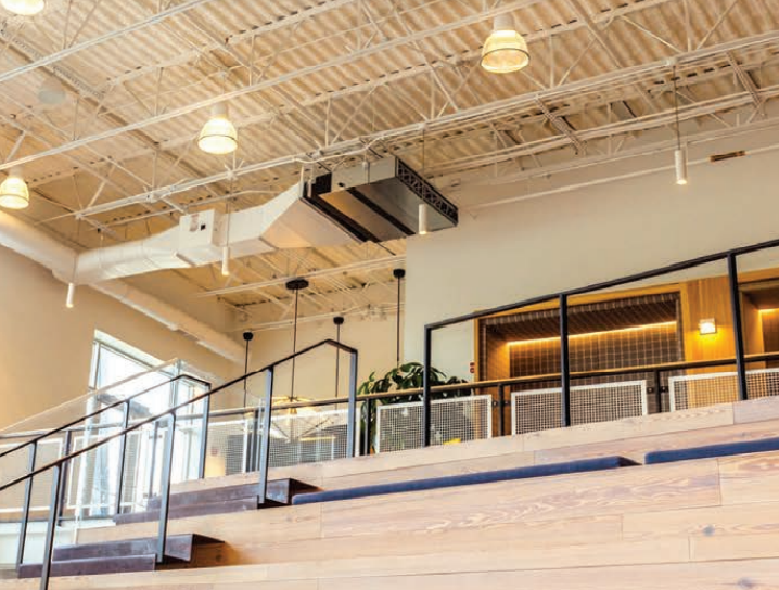 Modern interior with exposed ceiling, hanging lights, wooden bleacher seating, metal railings, and an upper level featuring a Mitsubishi PEAD indoor ceiling-concealed air handler for high energy efficiency.