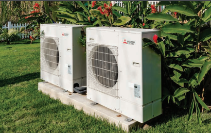 Two Mitsubishi Outdoor Single Zone Standard Condenser units (PUY, PUZ) are installed on a concrete slab in a garden with grass and tropical plants, designed for efficient cooling even in low ambient conditions.